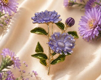 May include: A gold-tone metal brooch shaped like a cluster of purple flowers with green leaves. The brooch features two fully bloomed purple flowers and one small purple flower bud, all accented with vibrant green enamel leaves and stems.