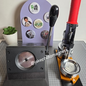 May include: A button maker machine with a red handle and a black base. A display board with several photo magnets is to the left. The magnets feature photos and the text "JAKE photo magnets". The Instagram handle @jakephotomagnets is visible.