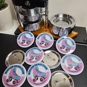 May include: A selection of round badges featuring a pink and blue roller skate design, with the text "ROLLER SKATE CLUB" and "PRINCESS ON SKATES". A button-making machine is visible in the background.
