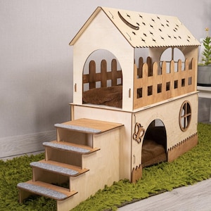 May include: A wooden cat house with a house-shaped design. It features a two-story structure with a staircase, a fenced balcony, and a decorative roof. The interior includes a brown cushion for comfort. The house is set on a green rug.