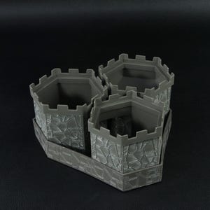 May include: A gray, castle-themed planter with three hexagonal pots connected by a rectangular base. The pots have a textured, stone-like appearance and crenelated tops. The planter is designed to hold multiple plants, adding a unique touch to any space.
