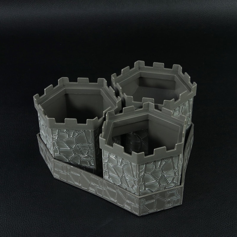 May include: A gray, castle-themed planter with three hexagonal pots connected by a rectangular base. The pots have a textured, stone-like appearance and crenelated tops. The planter is designed to hold multiple plants, adding a unique touch to any space.