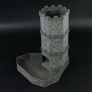 May include: A gray plastic dice tower and tray set. The tower has a stone-like texture and a hexagonal top. The tray is also gray and has a matching stone-like texture. The set is designed for tabletop gaming.
