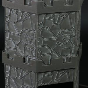 May include: Close-up of a gray, stone-textured structure with a fortress-like design. The surface is divided into irregular shapes, creating a rugged appearance. Dark gray bands encircle the structure, adding to its architectural detail.