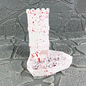 Könnte beinhalten: Ein weißer Würfelturm mit roten Spritzern, der einem Burgturm ähnelt, steht auf einem passenden Tablett. Mehrere weiße Würfel mit schwarzen Zahlen befinden sich im Tablett. Für Tischspiele.