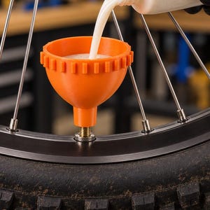 May include: An orange plastic funnel is attached to a bicycle tire valve stem. A translucent squeeze bottle is pouring a white liquid sealant into the funnel. The funnel has a ribbed outer edge and a threaded metal base connecting to the tire valve.