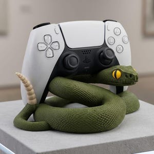 May include: A white and black video game controller resting on a coiled green rattlesnake figure. The snake has a textured body, a tan rattle at the tip of its tail, and large yellow eyes with black pupils. The controller features standard buttons.