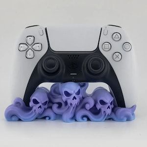 Può includere: Un controller per videogiochi bianco e nero appoggiato su un supporto viola chiaro. Il supporto presenta un design scolpito di tre teschi integrati in forme vorticose simili a nuvole che sostengono saldamente la base del controller.