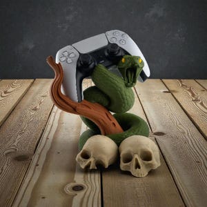 May include: A white and black video game controller is held by a green snake sculpture with an open mouth. The snake is coiled around a brown branch, with two skulls at the base. The background is a dark gray wall and a wooden surface.