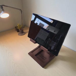 May include: A brown tablet stand with a tablet in place, set on a light wood desk. A silver desk lamp and small potted plants are also on the desk. The tablet screen reflects the room.