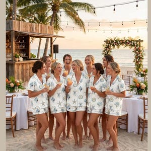 May include: A group of women on a beach, wearing matching white pyjama sets with a blue sea life pattern. They are laughing and holding drinks, with a bar and floral arch in the background. The scene is set at sunset.