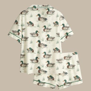 Puede incluir: Un conjunto de pijama de dos piezas con un fondo color crema con un patr&oacute;n repetido de patos nadando en un estanque. La parte superior de manga corta y los pantalones cortos tienen ribetes verdes.