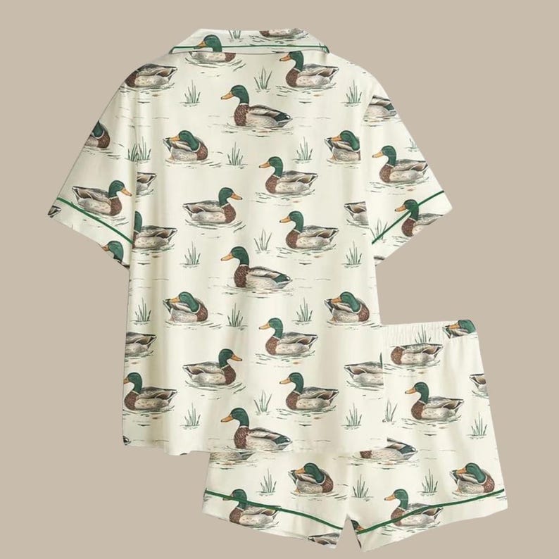 Puede incluir: Un conjunto de pijama de dos piezas con un fondo color crema con un patr&oacute;n repetido de patos nadando en un estanque. La parte superior de manga corta y los pantalones cortos tienen ribetes verdes.