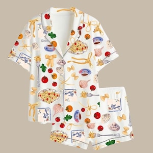 May include: White pajama set with a pasta-themed print. The short-sleeved top and shorts feature illustrations of pasta dishes, tomatoes, and bow-tie pasta. Accents include yellow bows and the text "Bon Appetit" on the shorts.