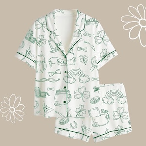 Może przedstawiać: Biały zestaw piżamy z zielonymi wykończeniami i nadrukiem w tematyce Dnia Świętego Patryka. Nadruk zawiera koniczyny, tęcze, garnki złota i słowo "lucky". Zestaw zawiera koszulę zapinaną na guziki i szorty.