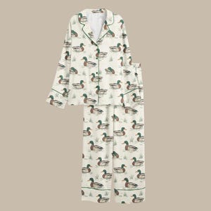 Puede incluir: Conjunto de pijama color crema con un estampado repetido de patos en un estanque. La parte superior de manga larga y los pantalones tienen ribetes verdes. Los patos son en tonos marrones, verdes y azules.
