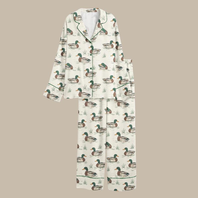 Puede incluir: Conjunto de pijama color crema con un estampado repetido de patos en un estanque. La parte superior de manga larga y los pantalones tienen ribetes verdes. Los patos son en tonos marrones, verdes y azules.