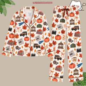 Puede incluir: Un conjunto de pijama de dos piezas con camisa de manga larga abotonada y pantalón a juego sobre un fondo crema. El estampado muestra ilustraciones de temática otoñal que incluyen calabazas naranjas, hojas de arce rojas, todoterrenos marrones, cenadores, libros y tazas de café.