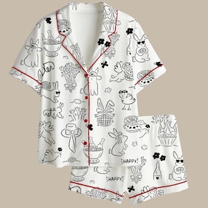 Peut inclure: Ensemble de pyjama blanc avec un motif noir et rouge. L'ensemble comprend une chemise boutonn&eacute;e &agrave; manches courtes et un short assorti. L'imprim&eacute; repr&eacute;sente des lapins, des fleurs, des g&acirc;teaux et le mot "HAPPY".
