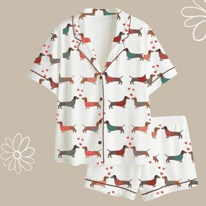 May include: White pyjama set featuring a repeating pattern of dachshund dogs in various plaid and polka dot outfits. The set includes a button-down shirt and shorts, both trimmed with brown piping.