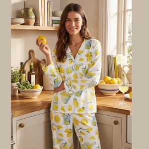 Könnte beinhalten: Weißes Pyjama-Set mit Zitronenmuster. Das langärmlige Oberteil und die Hose zeigen ein sich wiederholendes Muster aus gelben Zitronen und Limonadenflaschen. Die Frau im Bild hält eine Zitrone.