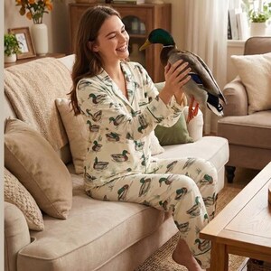 Puede incluir: Conjunto de pijama color crema con estampado de patos. La parte superior de manga larga y los pantalones presentan un dise&ntilde;o repetido de patos en el agua. El pijama tiene ribetes verdes y se usa en un sal&oacute;n.