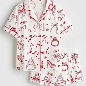 Op de afbeelding: Witte pyjamaset met rode rand en een herhalend patroon van illustraties met een bruiloftthema. De set bevat een overhemd met korte mouwen en bijpassende shorts. Het overhemd heeft de tekst "going to the chapel".