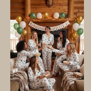 May include: A group of people in matching white pyjamas with a green tree pattern, celebrating a bachelorette party. The scene includes balloons, a banner that says "Bachelorette", and gifts.