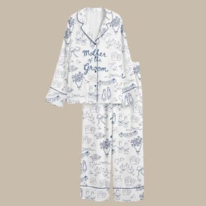 May include: White pajama set with a blue floral and wedding-themed pattern. The top features the text "Mother of the Groom" in blue script. The set includes a button-down top and matching pants, perfect for a wedding.