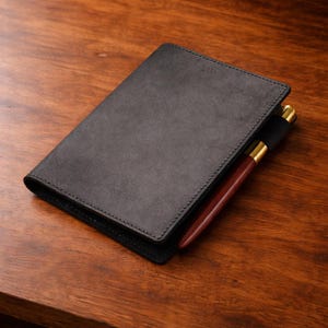 May include: A black leather journal with a pen holder and a wooden pen with gold accents. The journal is closed and has a stitched border. The pen is partially inserted into the journal's pen loop. The journal is on a wooden surface.