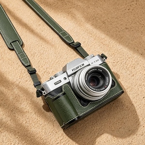 May include: A silver Fujifilm X-T30 camera with a green leather case and strap. The camera has a lens and is resting on a sandy surface. The strap is attached to the camera case with white stitching.