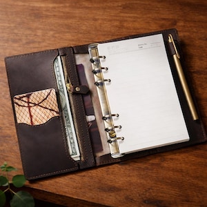 Könnte beinhalten: Offenes braunes Lederjournal mit linierten Notizblock, Stift und Kartenhalter. Das Journal hat eine Reißverschlusstasche mit Bargeld. Der Stift ist goldfarben. Das Journal liegt offen auf einer Holzoberfläche.