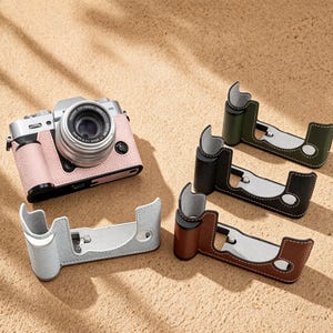 May include: A digital camera with a pink leather case sits next to four leather camera grips in various colors: gray, brown, black, and olive green. The grips have a silver base and white stitching, resting on a sandy surface.