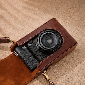 Könnte beinhalten: Eine braune Leder-Kameratasche mit einer schwarzen Kamera im Inneren. Die Tasche hat einen Schulterriemen und einen Schnallenverschluss. Das Kameraobjektiv ist sichtbar, und die Tasche scheint aus hochwertigem Leder gefertigt zu sein.