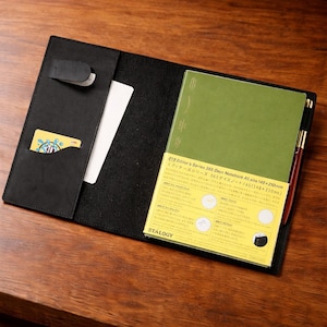 Puede incluir: Funda de cuaderno de cuero negro con un cuaderno verde y un bolígrafo rojo. La funda tiene un bolsillo con una tarjeta amarilla y una tarjeta blanca. El cuaderno tiene el texto "018 Editor's Series 365 Days Notebook A5 size 148x210mm". El cuaderno mide 14,8 cm x 21 cm.