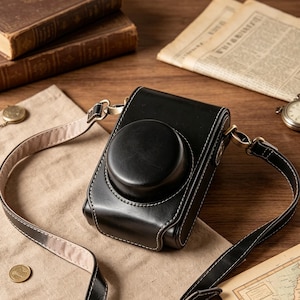 Puede incluir: Una funda negra para cámara con cuerpo rectangular estructurado y tapa circular para el objetivo. La funda presenta costuras blancas en contraste, herrajes metálicos y una correa ajustable para el hombro con forro interior claro.