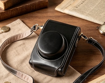 May include: A black camera case with a structured rectangular body and a circular lens cover. The case features white contrast stitching, metallic hardware, and an adjustable shoulder strap with a light-colored interior lining.