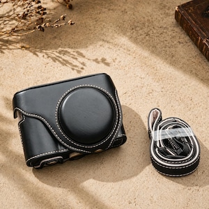 May include: Black leather camera case with white stitching and a matching strap. The case has a circular lens opening and a snap closure. The strap is black and white with a padded shoulder rest. The case is designed to protect a digital camera.