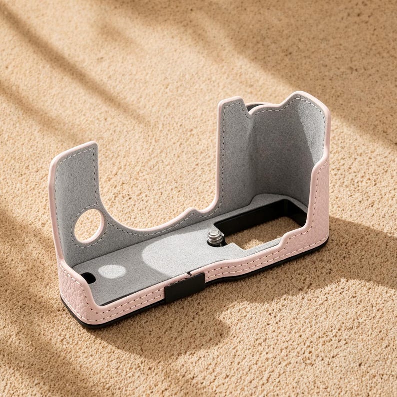 May include: A pink leather camera case with a gray interior. The case has a curved design with a cutout for the lens and a black strap. The case is sitting on a sandy surface.