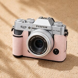 May include: A silver Fujifilm X-T30 camera with a black and pink leather case. The camera lens is labeled "FUJINON ASPHERICAL LENS". The camera is on a sandy surface.