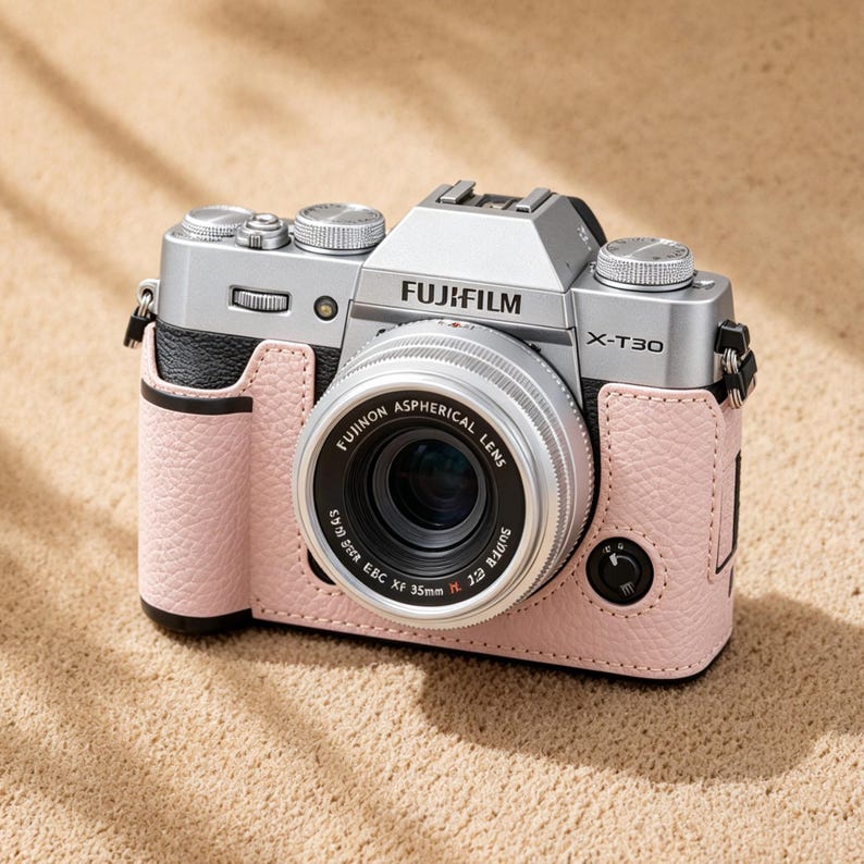 May include: A silver Fujifilm X-T30 camera with a black and pink leather case. The camera lens is labeled "FUJINON ASPHERICAL LENS". The camera is on a sandy surface.