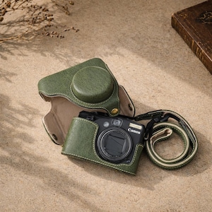 Può includere: Una custodia per fotocamera in pelle verde con un copriobiettivo e un cinturino abbinati. La custodia è aperta, rivelando una fotocamera Canon nera. La custodia e il cinturino hanno cuciture bianche. L'obiettivo della fotocamera è visibile.