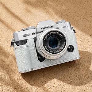 May include: A silver Fujifilm X-T30 camera with a black lens, encased in a light gray leather case. The camera is positioned on a sandy surface, with the Fujifilm logo clearly visible. The lens reads "Fujinon Aspherical Lens".