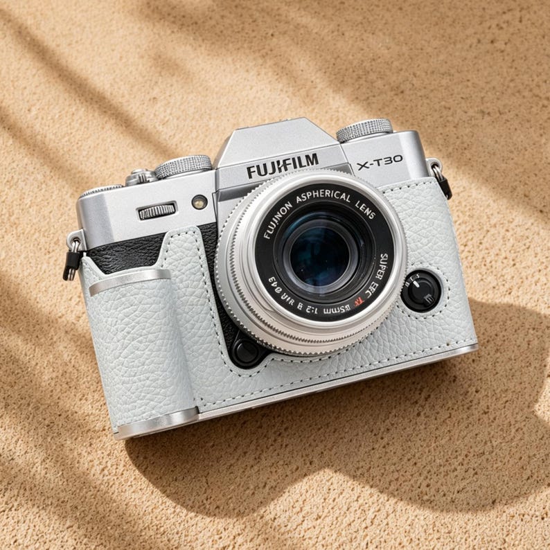 May include: A silver Fujifilm X-T30 camera with a black lens, encased in a light gray leather case. The camera is positioned on a sandy surface, with the Fujifilm logo clearly visible. The lens reads "Fujinon Aspherical Lens".