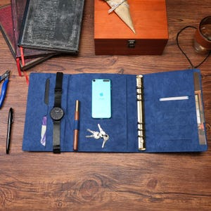 Op de afbeelding: Een blauwe organizer van kunstleer met meerdere binnenvakken, een goudkleurig mechanisme met zes ringen, een zwart polshorloge, een bruine pen, een lichtblauwe smartphone en een set zilveren sleutels op het oppervlak.