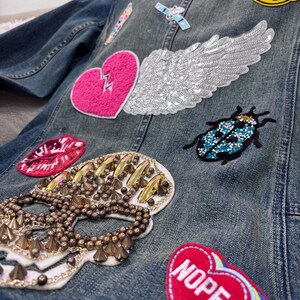 Puede incluir: Chaqueta vaquera azul adornada con varios parches, incluyendo un corazón rosa con un rayo, un ala con lentejuelas, una calavera y un corazón rojo con la palabra "NOPE". Otros parches incluyen una carita sonriente y un escarabajo azul.