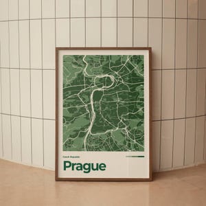 Op de afbeelding: Een ingelijste kaartprint van Praag, Tsjechië, in tinten groen en wit. De stadskaart is gedetailleerd met wegen en bezienswaardigheden. De print zit in een bruine houten lijst en het woord "Prague" is groen onderaan gedrukt.