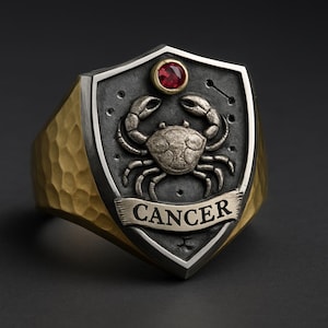 Pode incluir: Um anel de prata e ouro com o signo do zodíaco de Câncer. O anel tem uma face em forma de escudo com um design de caranguejo, uma pedra preciosa vermelha e a palavra "CANCER" em uma faixa. A banda tem uma textura martelada.