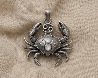 May include: A silver-toned metal pendant shaped like a crab, featuring a textured shell, two raised claws, and a small circular white stone set in the center. Above the crab is the Cancer zodiac symbol, and a bail is attached at the very top.
