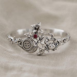 May include: A silver-toned bracelet featuring a floral and crab design. The bracelet includes a red gemstone and the Cancer zodiac symbol. The design is intricate and detailed, with a polished finish.
